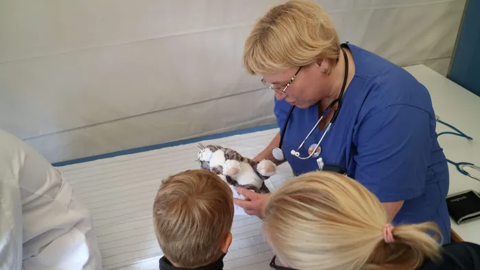 Eine Frau in medizinischer Kleidung untersucht ein Kuscheltier auf einem Tisch, während zwei Kinder zusehen.