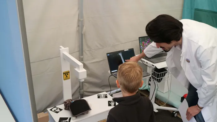 Ein Mann im weißen Kittel zeigt einem Jungen auf einem Laptop ein Röntgenbild. Auf dem Tisch liegt ein Kuscheltier.