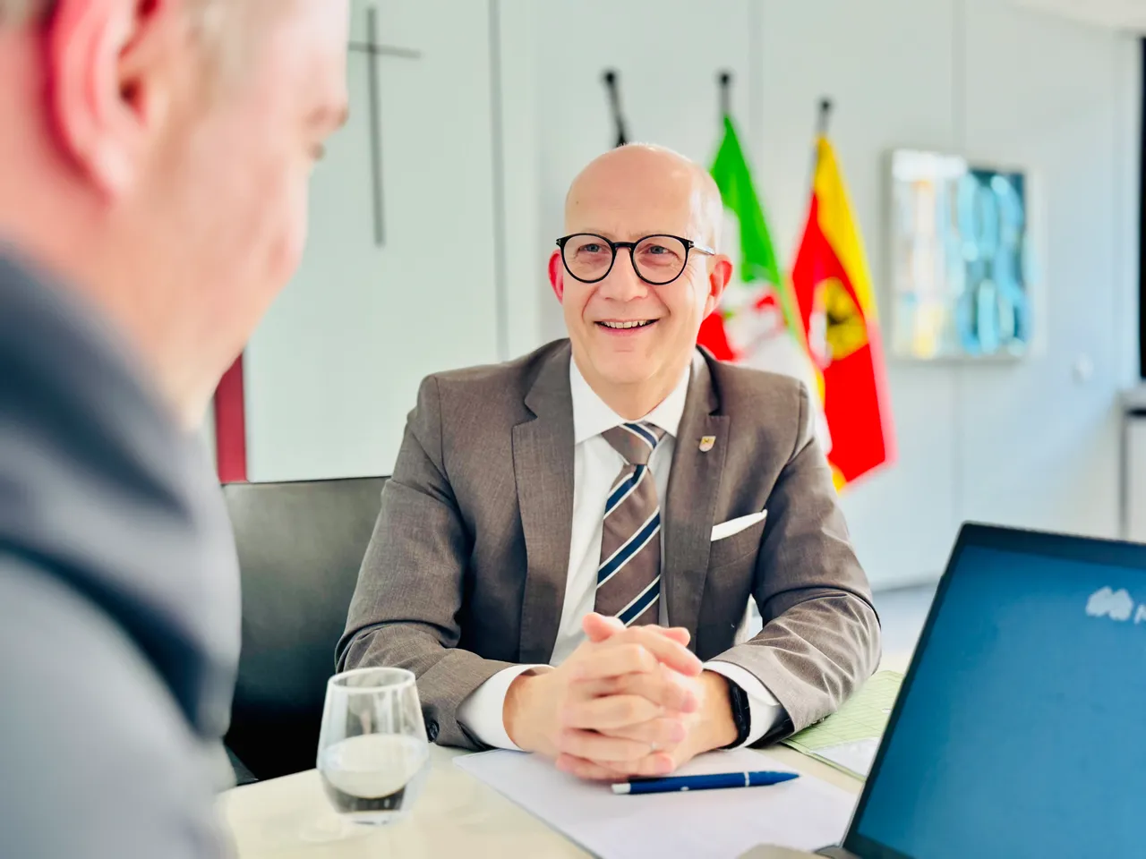 Landrat Ralf Schwarzkopf mit Brille und Anzug lächelt während eines Interviews. Im Hintergrund sind Flaggen zu sehen.