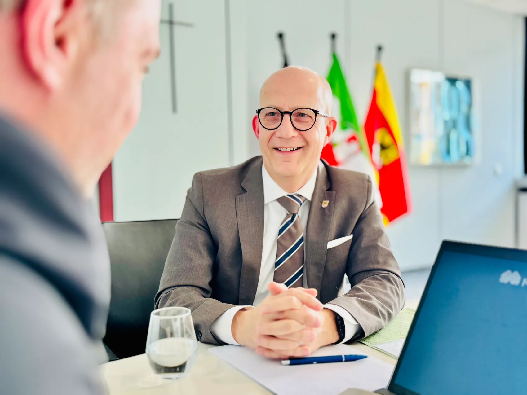 Landrat Ralf Schwarzkopf mit Brille und Anzug lächelt während eines Interviews. Im Hintergrund sind Flaggen zu sehen.