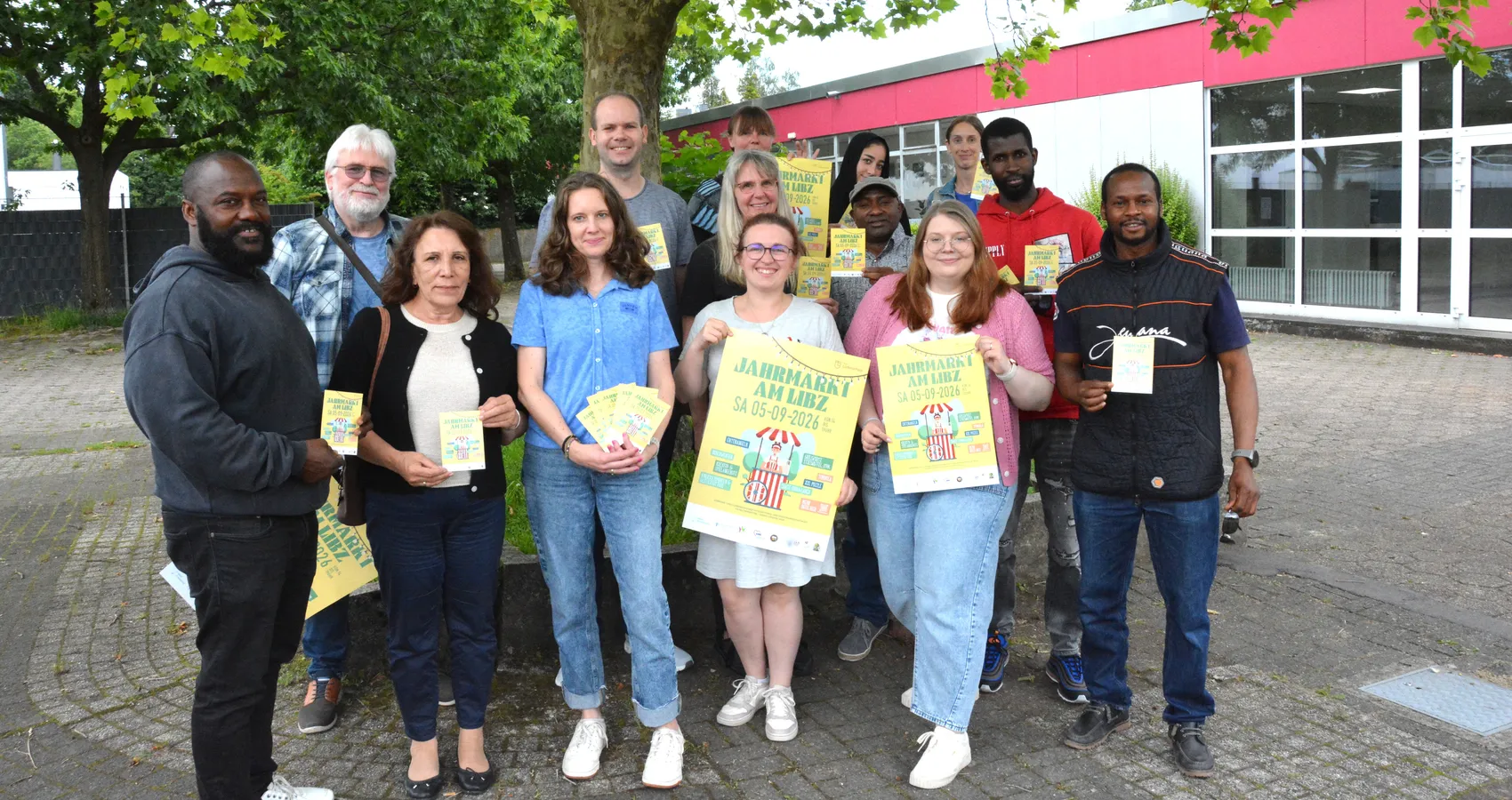 Eine Gruppe von etwa 15 Personen steht lächelnd vor einem roten Gebäude mit großen Fenstern. Viele halten gelbe Flyer mit der Aufschrift „Jahrmarkt am LIBZ Sa 05.09.2026“ in die Kamera.