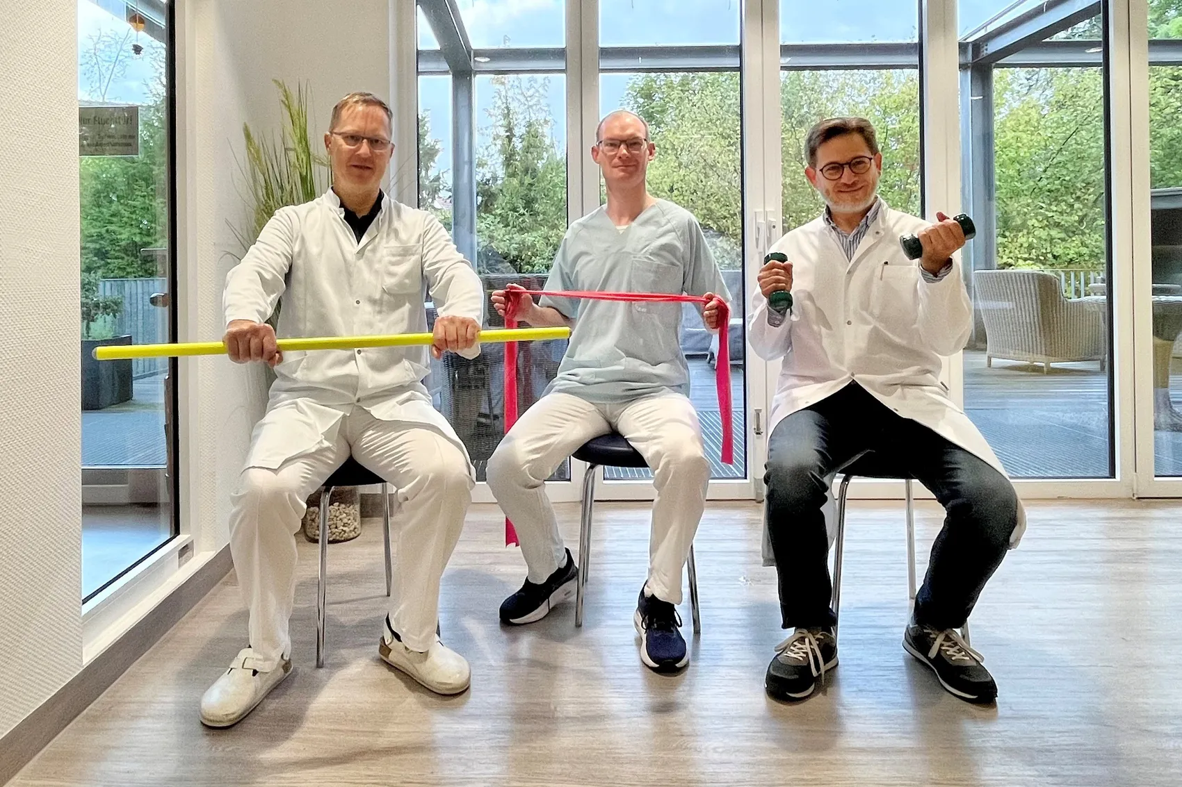 Drei Männer in medizinischer Kleidung sitzen in einem hellen Raum und halten einen gelben Stab, ein rosa Theraband und Hanteln.