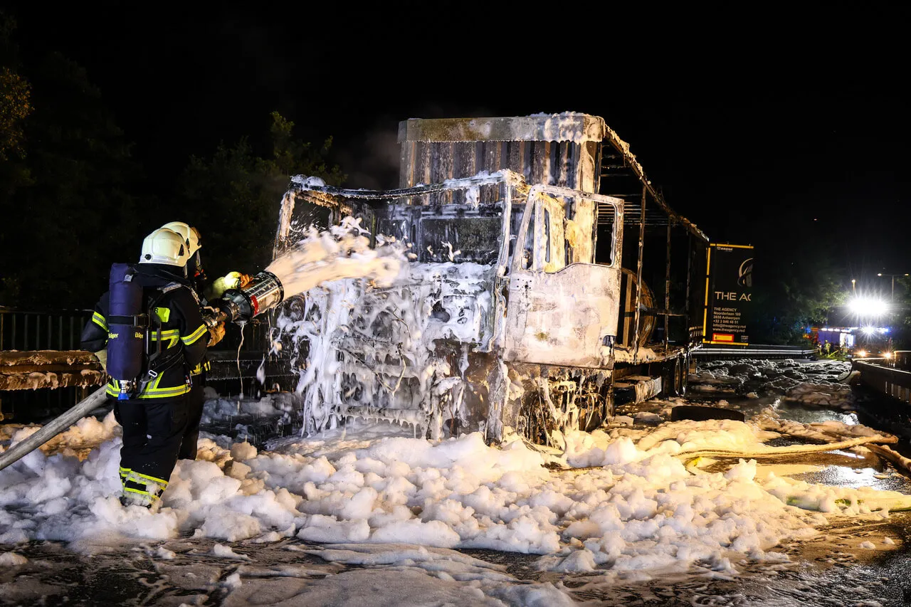 Ein Feuerwehrmann löscht mit einem Schlauch die Überreste eines ausgebrannten Lastwagens, der von Schaum bedeckt ist. Im Hintergrund ist ein beleuchtetes Gebäude mit der Aufschrift „THE AK“ zu sehen.