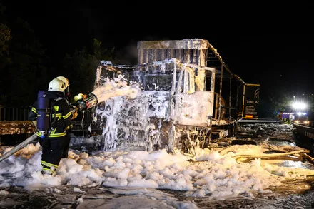 Ein Feuerwehrmann löscht mit einem Schlauch die Überreste eines ausgebrannten Lastwagens, der von Schaum bedeckt ist. Im Hintergrund ist ein beleuchtetes Gebäude mit der Aufschrift „THE AK“ zu sehen.
