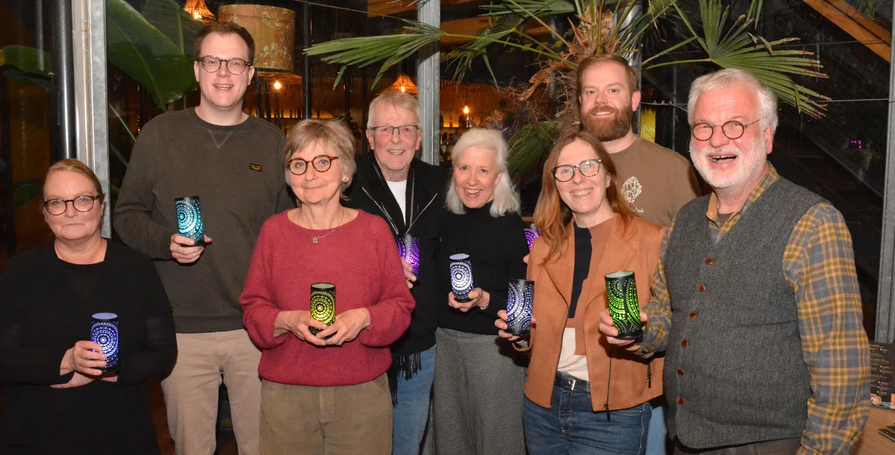 Sieben Personen halten bunte, leuchtende Gläser in der Hand, umgeben von Palmen und Pflanzen. Im Hintergrund sind weitere Personen und Lichtquellen erkennbar.