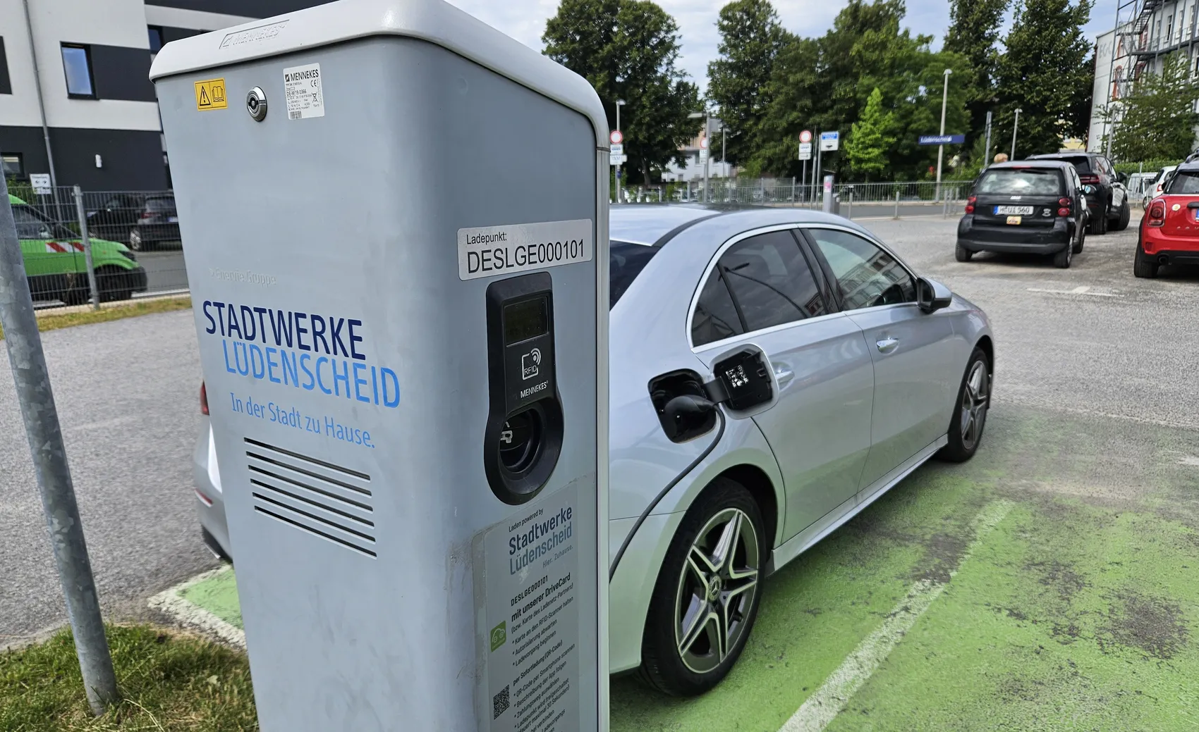 Ein silberner BMW i3 ist an einer blauen Ladestation der Stadtwerke Lüdenscheid angeschlossen. Die Station steht auf einem Parkplatz neben einer Straße.