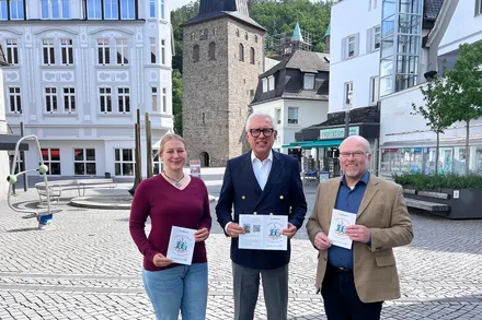 Drei Personen halten Informationsmaterial für einen Sponsorenlauf zur Sanierung der Christuskirche. Im Hintergrund ist die Kirche mit ihren Flankentürmchen zu sehen.