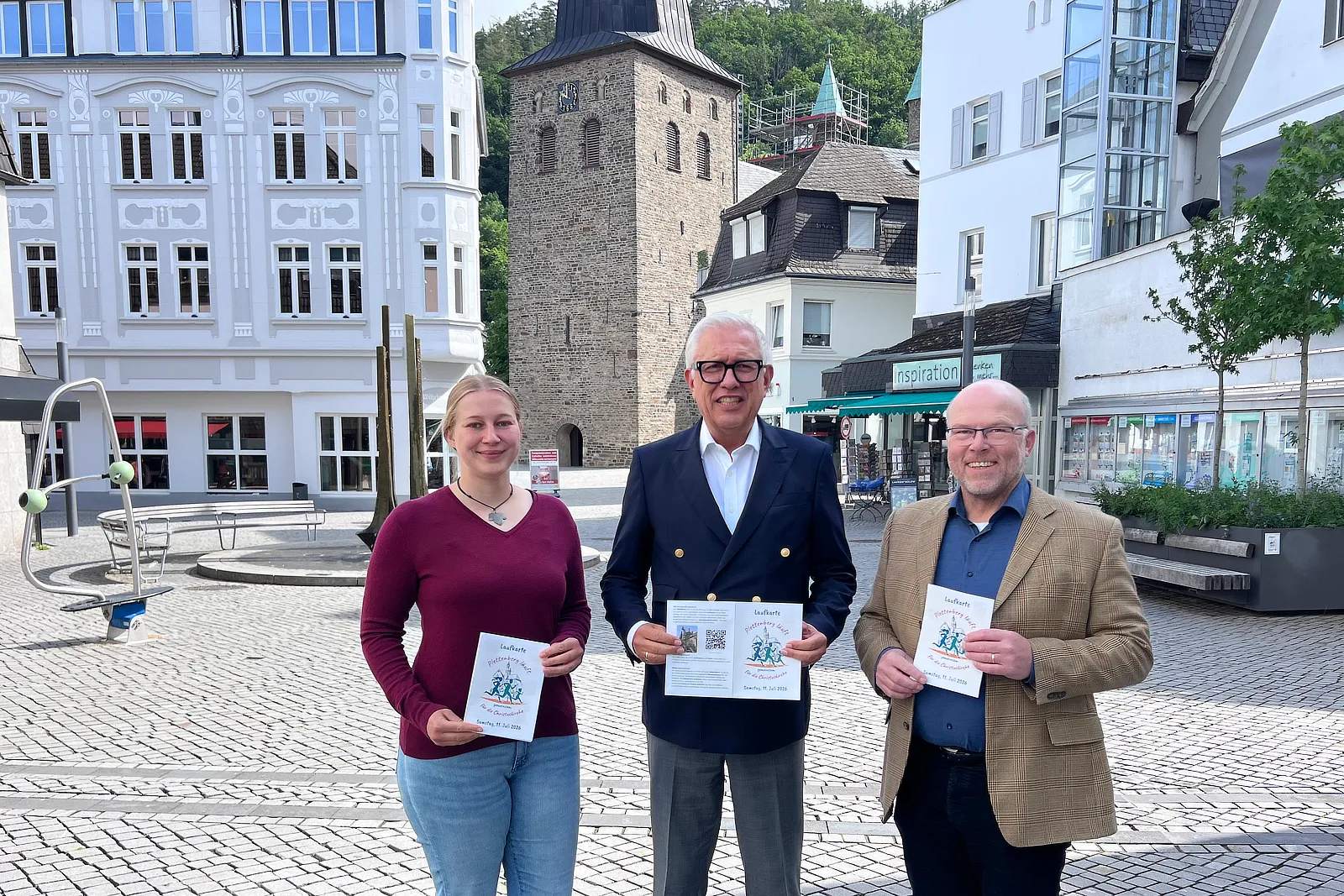 Drei Personen halten Informationsmaterial für einen Sponsorenlauf zur Sanierung der Christuskirche. Im Hintergrund ist die Kirche mit ihren Flankentürmchen zu sehen.