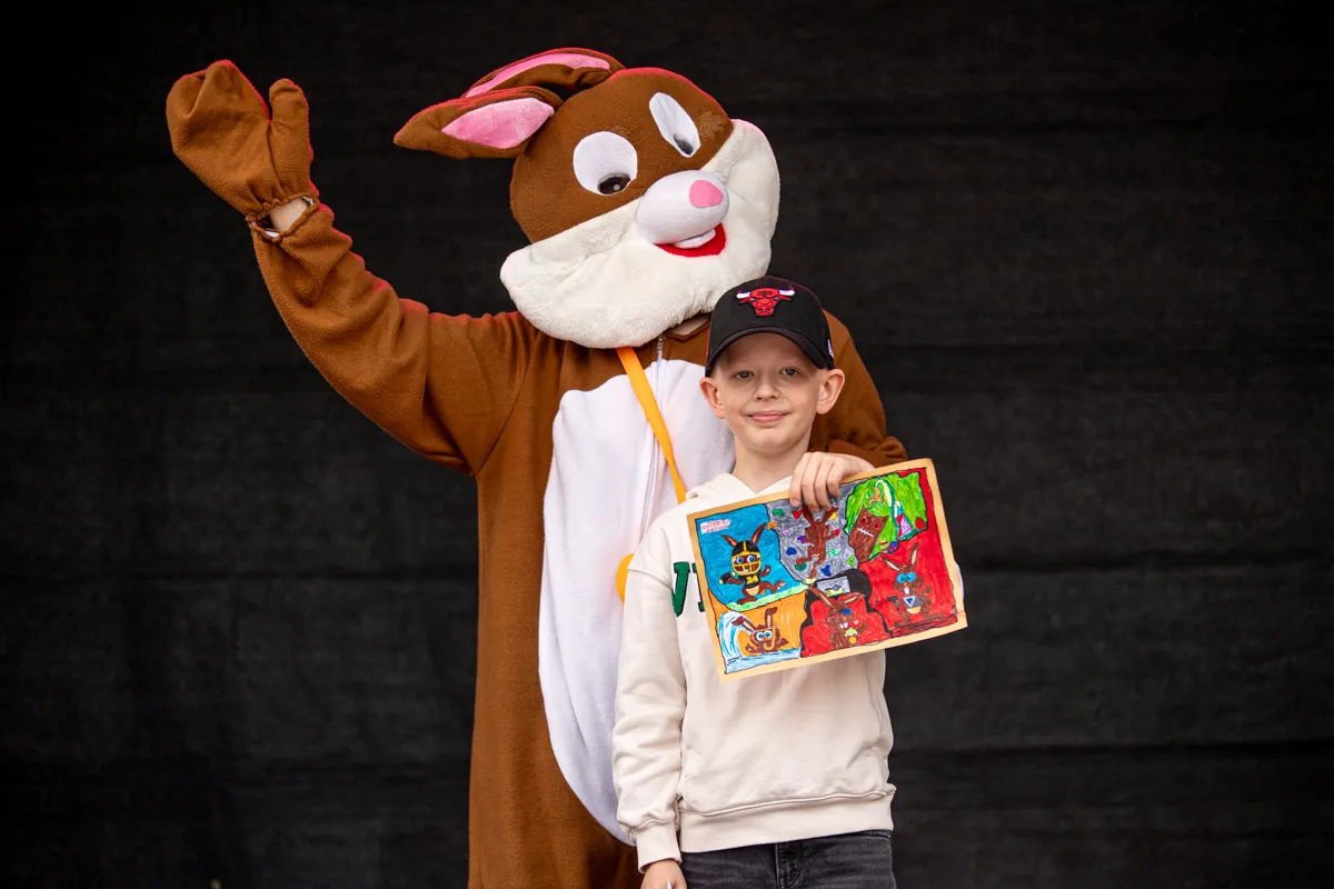 Ein Kind mit roter Baseballkappe hält ein buntes Bild vor sich, während ein großer Hasen-Kostümträger mit hochgehobener Pfote daneben steht.
