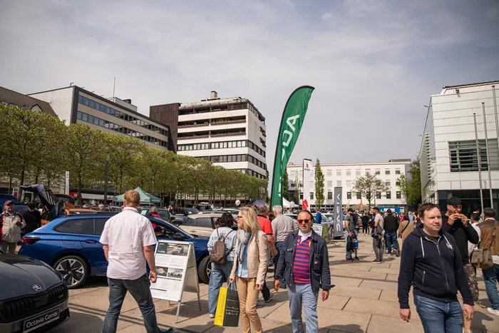 Menschen auf einem Stadtplatz mit Autos und Ständen, im Hintergrund moderne Bürogebäude und eine grüne Flagge mit dem Schriftzug „LDA“.
