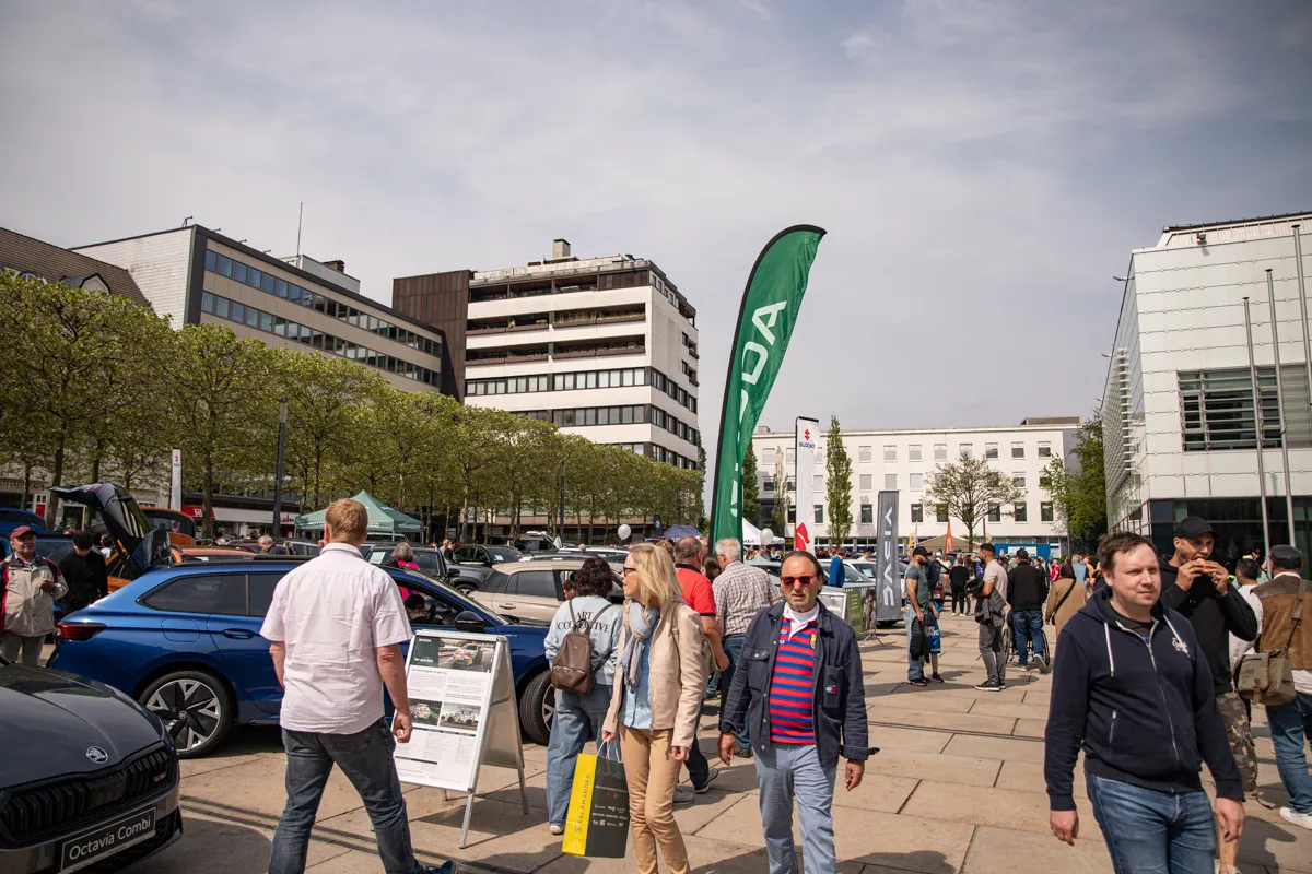 Menschen auf einem Stadtplatz mit Autos und Ständen, im Hintergrund moderne Bürogebäude und eine grüne Flagge mit dem Schriftzug „LDA“.