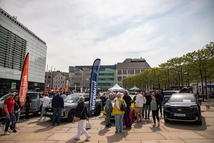 Menschengruppen betrachten auf einem Stadtplatz ausgestellte Ford-Fahrzeuge. Im Hintergrund sind Gebäude und weitere Besucher zu sehen.