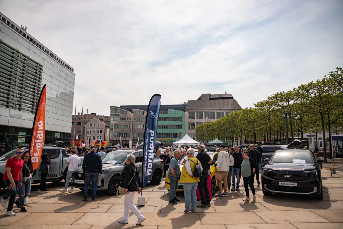 Menschengruppen betrachten auf einem Stadtplatz ausgestellte Ford-Fahrzeuge. Im Hintergrund sind Gebäude und weitere Besucher zu sehen.