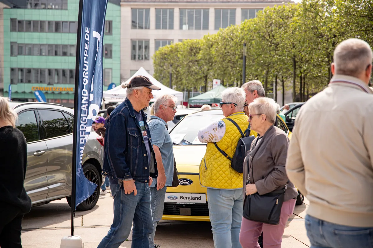 Mehrere Menschen stehen und unterhalten sich auf einem Stadtplatz vor einem gelben Ford und einer blauen Flagge mit der Aufschrift „will-bergland-gruppe.de“.