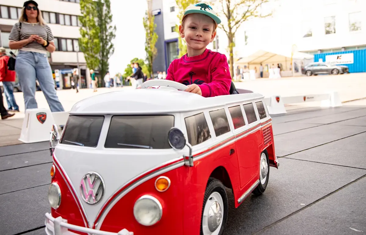 Ein Kind sitzt lächelnd am Steuer eines roten Spielzeug-VW-Busses im Freien, während im Hintergrund Menschen und Gebäude zu sehen sind.