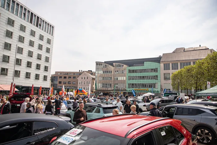 Menschenmengen und Autos füllen einen Stadtplatz an einem sonnigen Tag während des FIDLI-Festivals. Im Hintergrund sind moderne Gebäude und Geschäfte zu sehen.