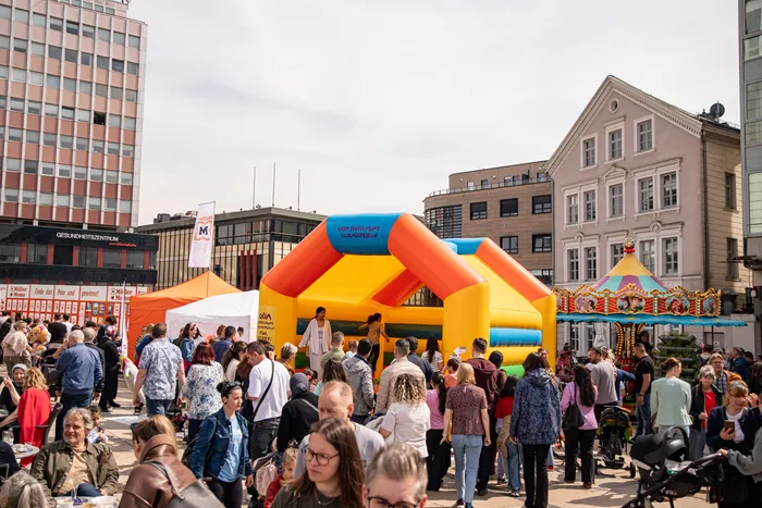 Menschen drängen sich auf einem Stadtplatz um eine große, farbenfrohe Hüpfburg. Im Hintergrund sind Gebäude und weitere Buden eines Stadtfestes zu sehen.
