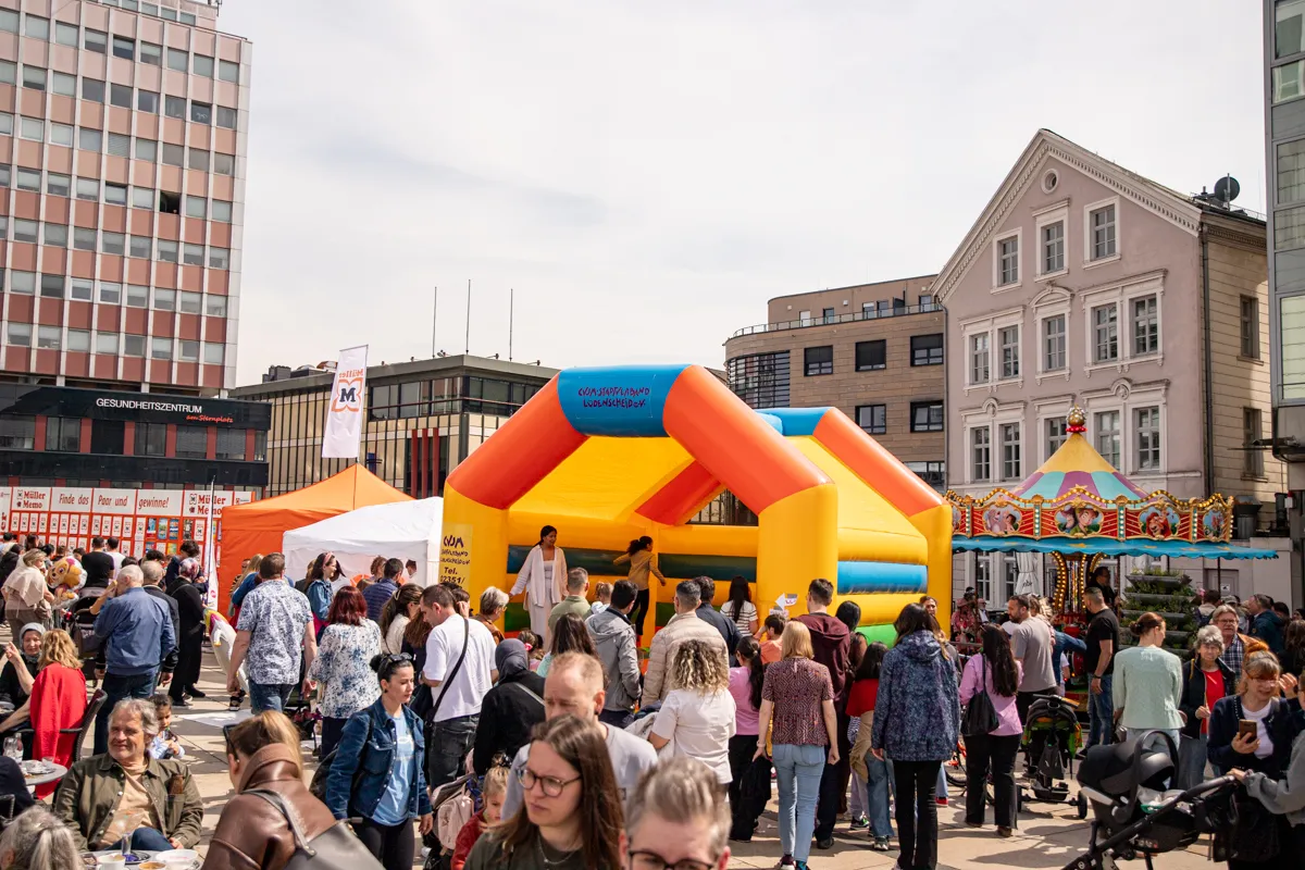 Menschen drängen sich auf einem Stadtplatz um eine große, farbenfrohe Hüpfburg. Im Hintergrund sind Gebäude und weitere Buden eines Stadtfestes zu sehen.