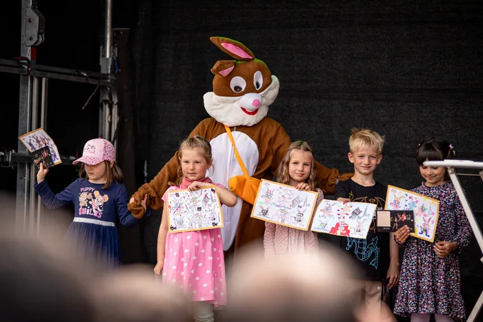 Fünf Kinder halten bemalte Osterkarten vor sich, während ein Osterhase im Hintergrund steht. Sie stehen auf einer Bühne vor dunklem Hintergrund.