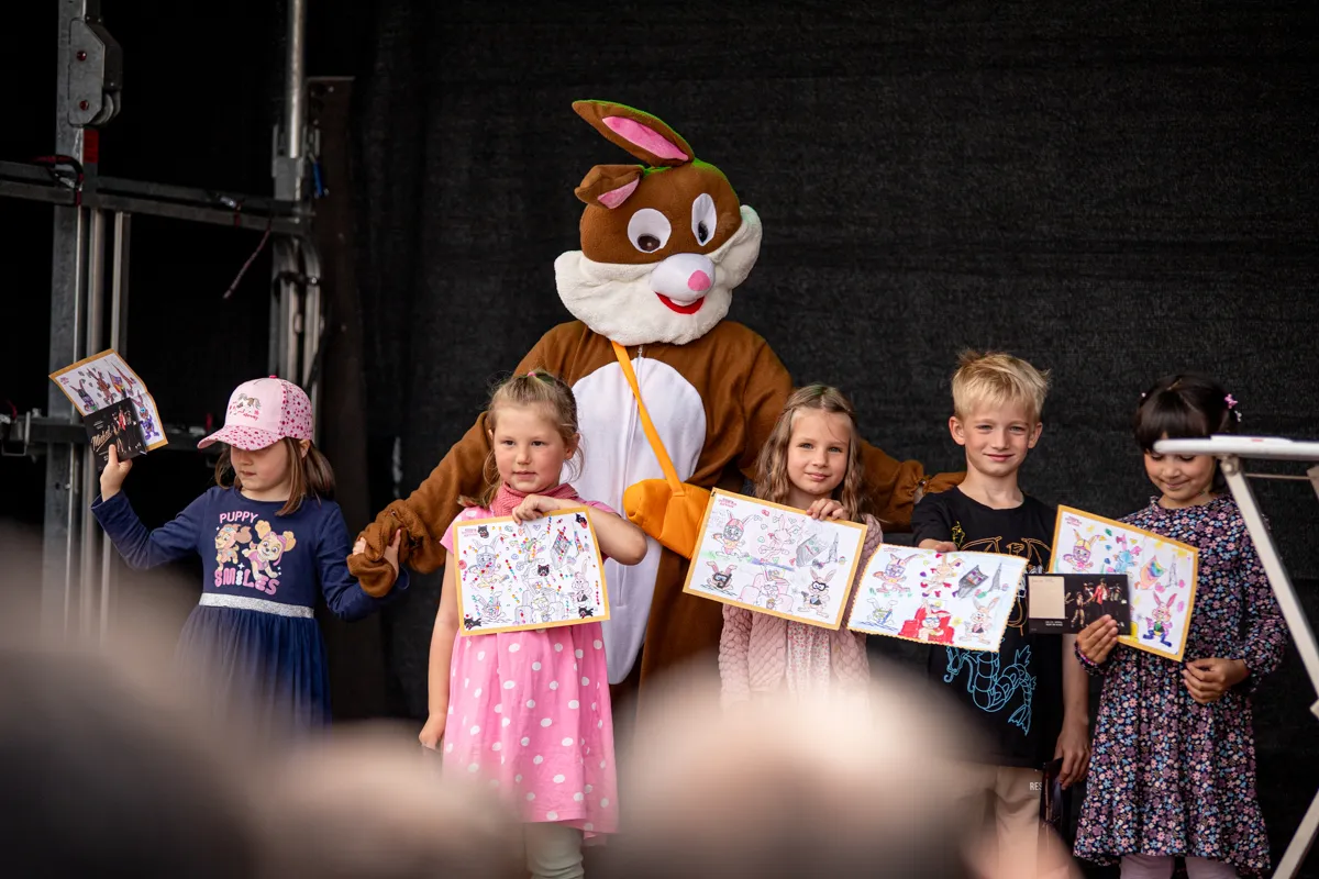 Fünf Kinder halten bemalte Osterkarten vor sich, während ein Osterhase im Hintergrund steht. Sie stehen auf einer Bühne vor dunklem Hintergrund.