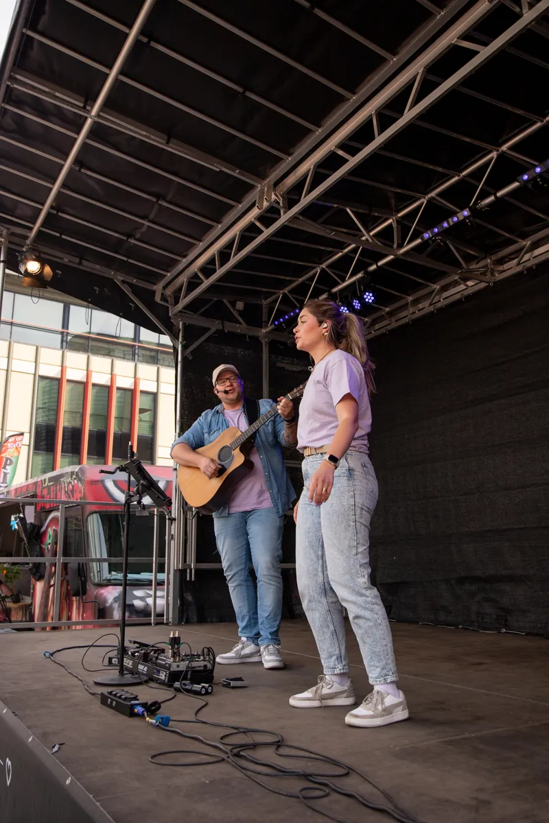 Zwei junge Erwachsene spielen auf einer Bühne im Freien. Die Frau singt in ein Mikrofon, während der Mann eine Akustikgitarre spielt.