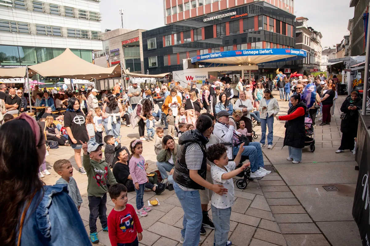 Große Menschenmenge auf einem Stadtplatz, viele Kinder stehen im Vordergrund und schauen einem Mann zu, der eine Seifenblase macht. Im Hintergrund Geschäfte und Pavillons.