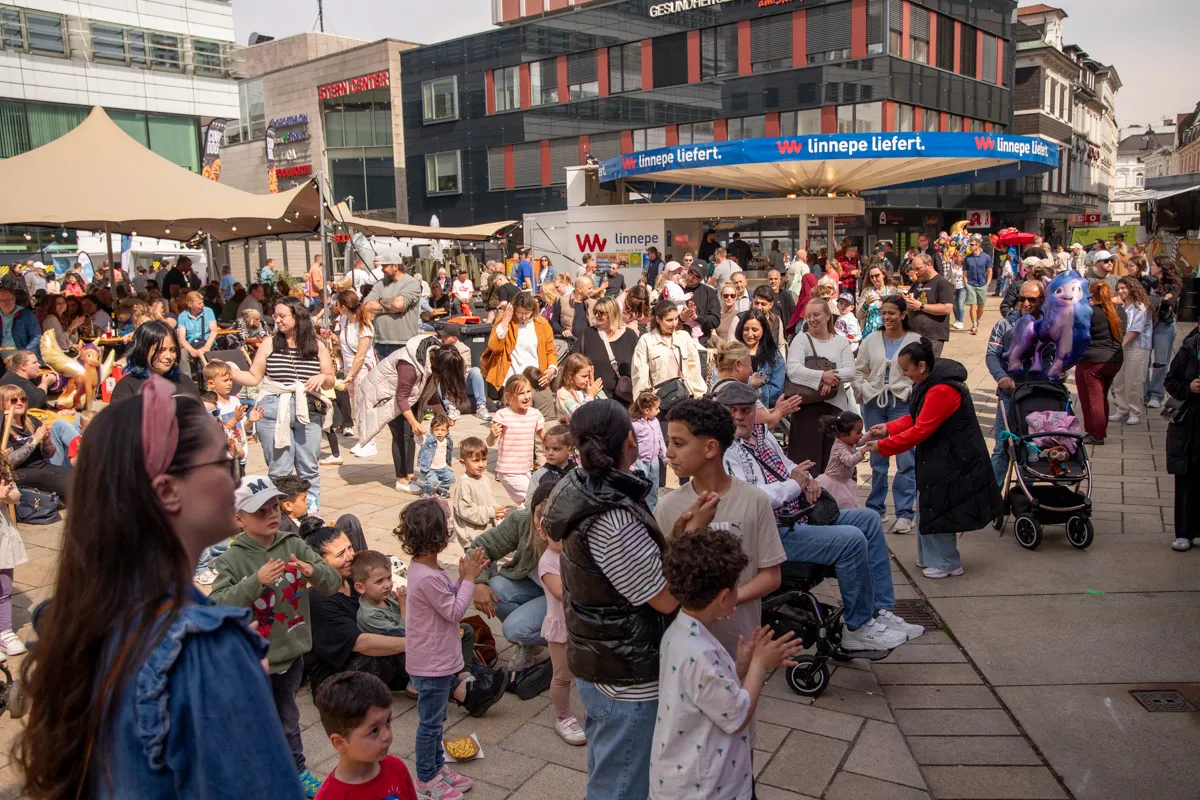 Große Menschenmenge auf einem Stadtplatz, viele Kinder sitzen und stehen im Vordergrund und schauen in Richtung einer Bühne oder eines Auftritts. Im Hintergrund sind Geschäfte und ein Banner mit der Aufschrift „linnepe liefert“ zu sehen.