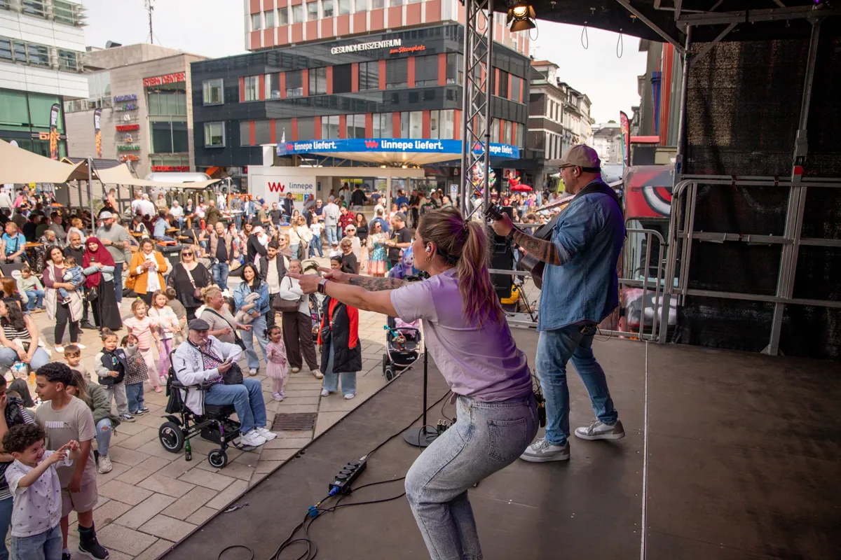 Eine Frau mit violetten Haaren gestikuliert auf einer Bühne, während ein Mann mit Gitarre neben ihr steht. Im Hintergrund eine große Menschenmenge und Gebäude in einer belebten Innenstadt.