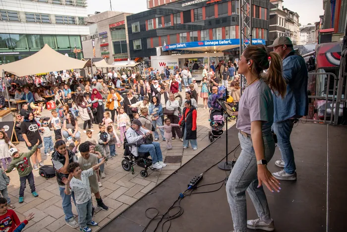 Zwei Personen stehen auf einer Bühne und performen vor einer großen Menschenmenge in einer belebten Innenstadt. Im Vordergrund tanzen Kinder, während sich im Hintergrund Geschäfte und Gebäude erstrecken.