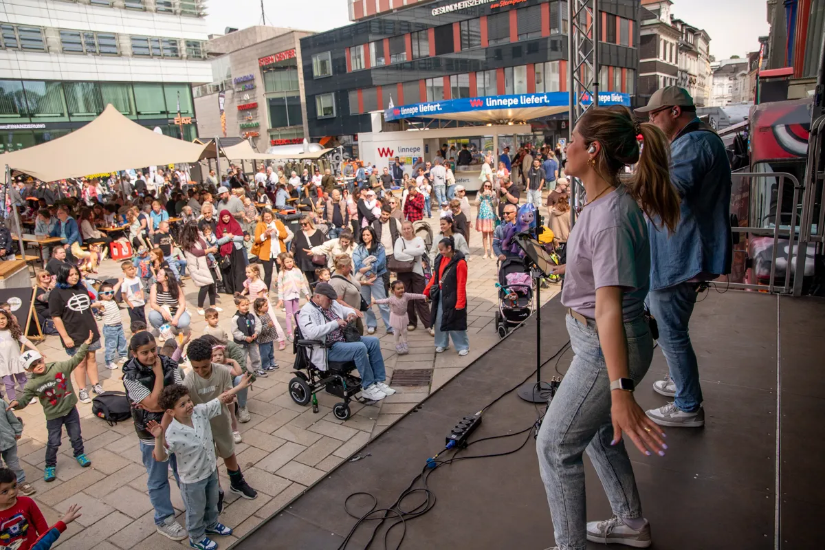 Zwei Personen stehen auf einer Bühne und performen vor einer großen Menschenmenge in einer belebten Innenstadt. Im Vordergrund tanzen Kinder, während sich im Hintergrund Geschäfte und Gebäude erstrecken.