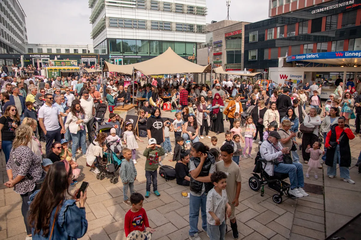 Volksfest mit vielen Besuchern auf einem Stadtplatz. Im Hintergrund moderne Gebäude und Geschäfte.