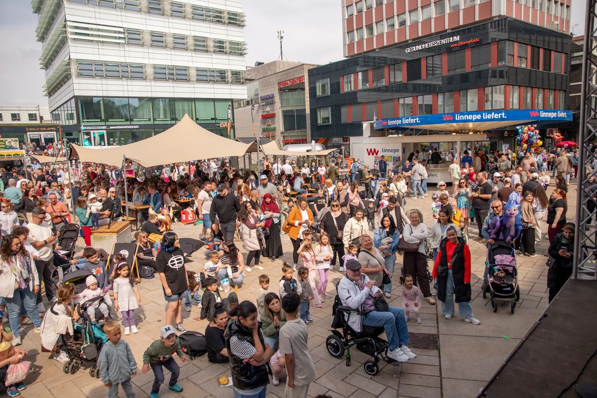 Volksfest mit vielen Besuchern auf einem Stadtplatz, umgeben von modernen Gebäuden und Geschäften. Im Hintergrund ein Gebäude mit der Aufschrift „GESUNDHEITSZENTRUM“.