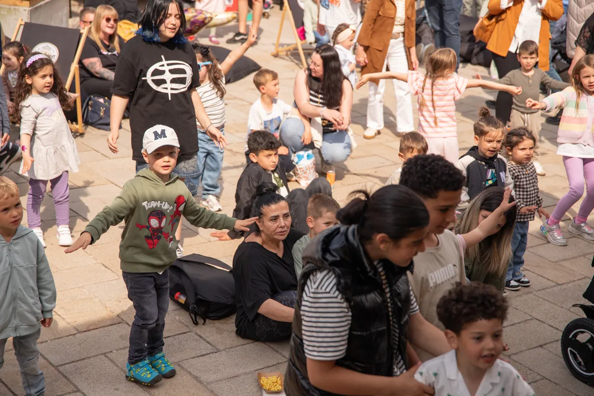 Kinder tanzen und spielen auf einem Stadtplatz, während Erwachsene zusehen. Im Hintergrund ist eine Frau mit einem schwarzen Oberteil und einer Mütze zu sehen.