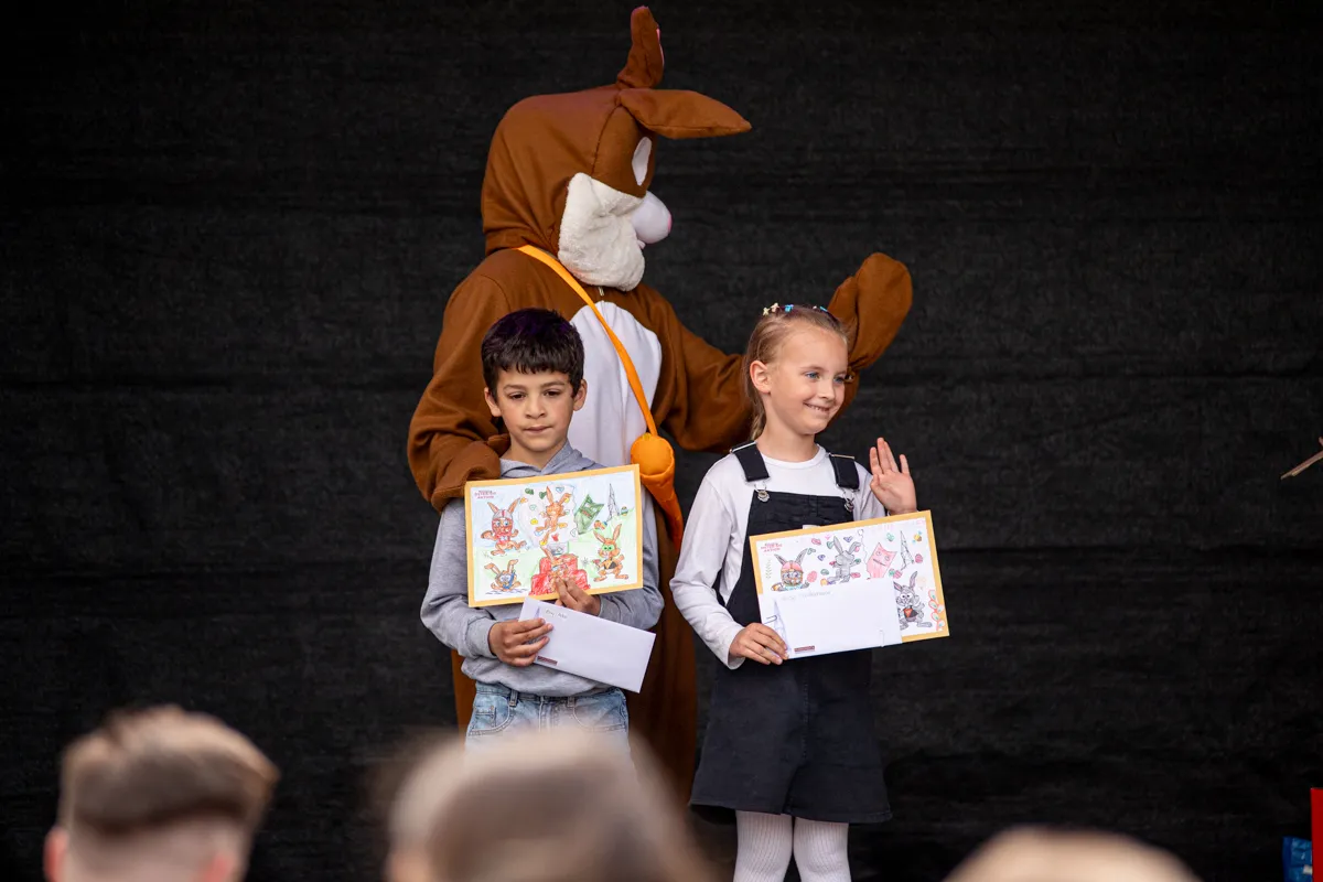 Zwei Kinder halten jeweils ein farbiges Bild und eine Urkunde auf einer Bühne. Im Hintergrund steht eine große Känguru-Figur.