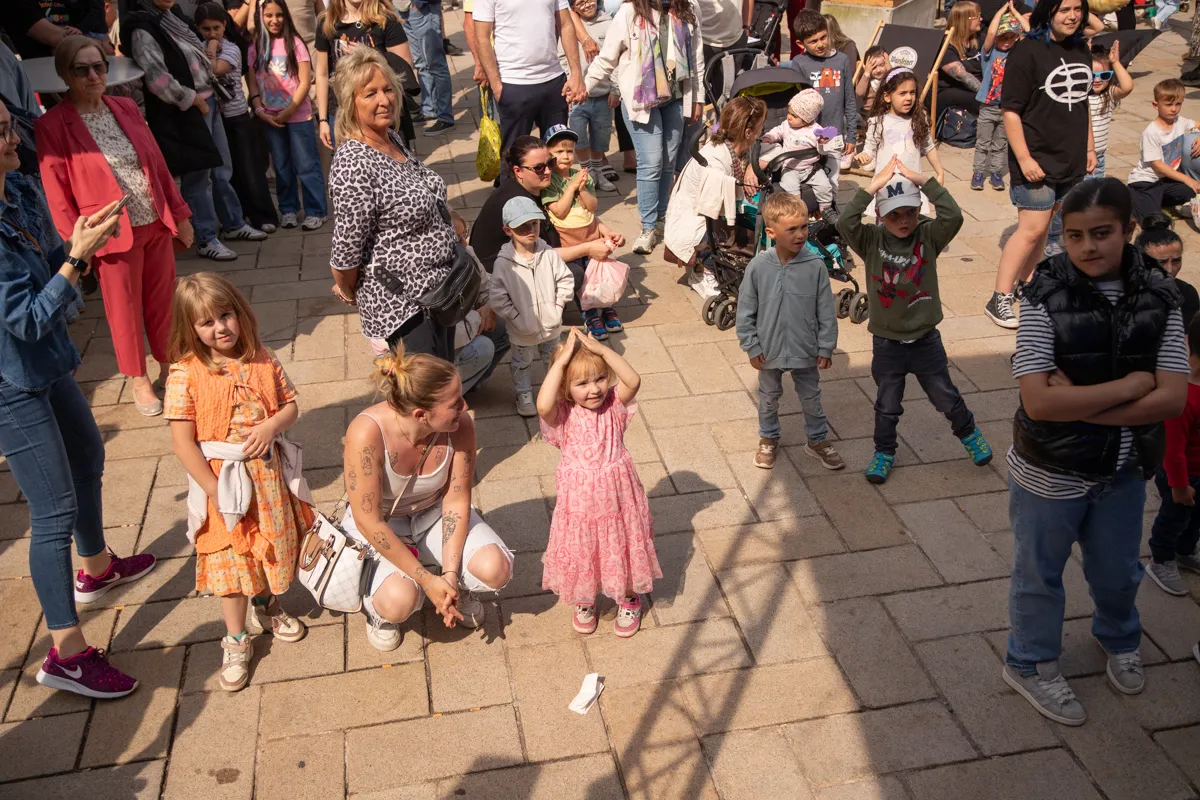 Eine Gruppe von Menschen, darunter viele Kinder, beobachtet eine Aktion auf einem gepflasterten Platz im Freien. Einige Kinder haben die Arme erhoben, während eine Frau in der Mitte hockt und etwas aufhebt.
