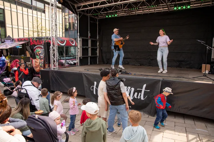 Ein Gitarrist und eine Sängerin treten auf einer Bühne vor einem Publikum mit Kindern auf. Im Hintergrund ein Stand mit Speisen und Getränken.