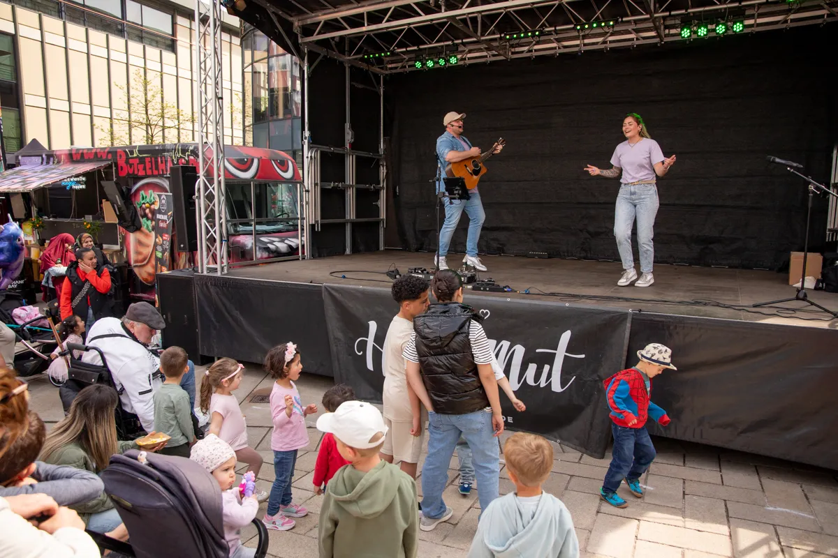 Ein Gitarrist und eine Sängerin treten auf einer Bühne vor einem Publikum mit Kindern auf. Im Hintergrund ein Stand mit Speisen und Getränken.