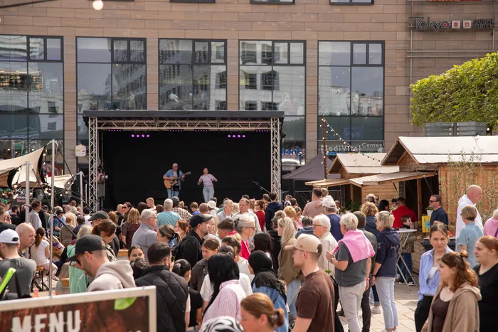 Musiker spielt Gitarre vor großem Publikum auf einem Stadtfest, im Hintergrund Geschäfte und Marktstände.