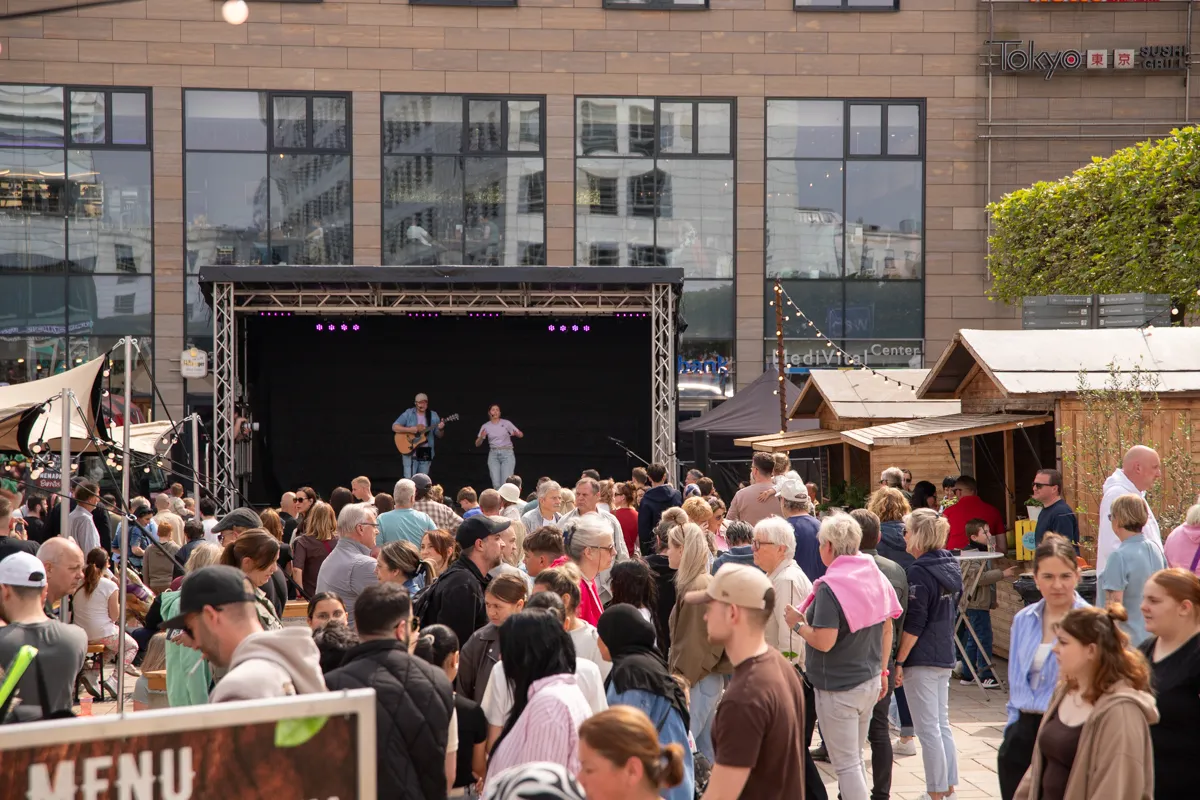 Musiker spielt Gitarre vor großem Publikum auf einem Stadtfest, im Hintergrund Geschäfte und Marktstände.