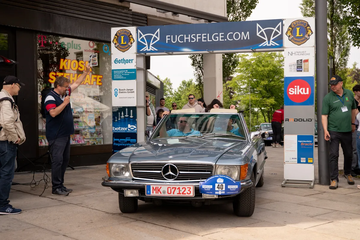 Silberner Mercedes SL mit geöffnetem Verdeck steht vor einem Kiosk und wird von Menschen betrachtet. Im Hintergrund sind Werbebanner und weitere Personen zu sehen.