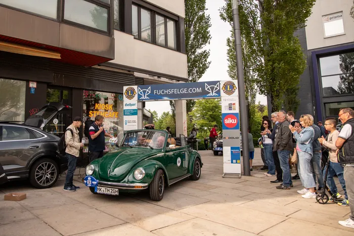 Ein dunkelgrüner VW Käfer steht im Mittelpunkt einer Menschenmenge vor Geschäften in einer Fußgängerzone. Im Hintergrund sind Werbebanner und ein grauer Tesla zu sehen.