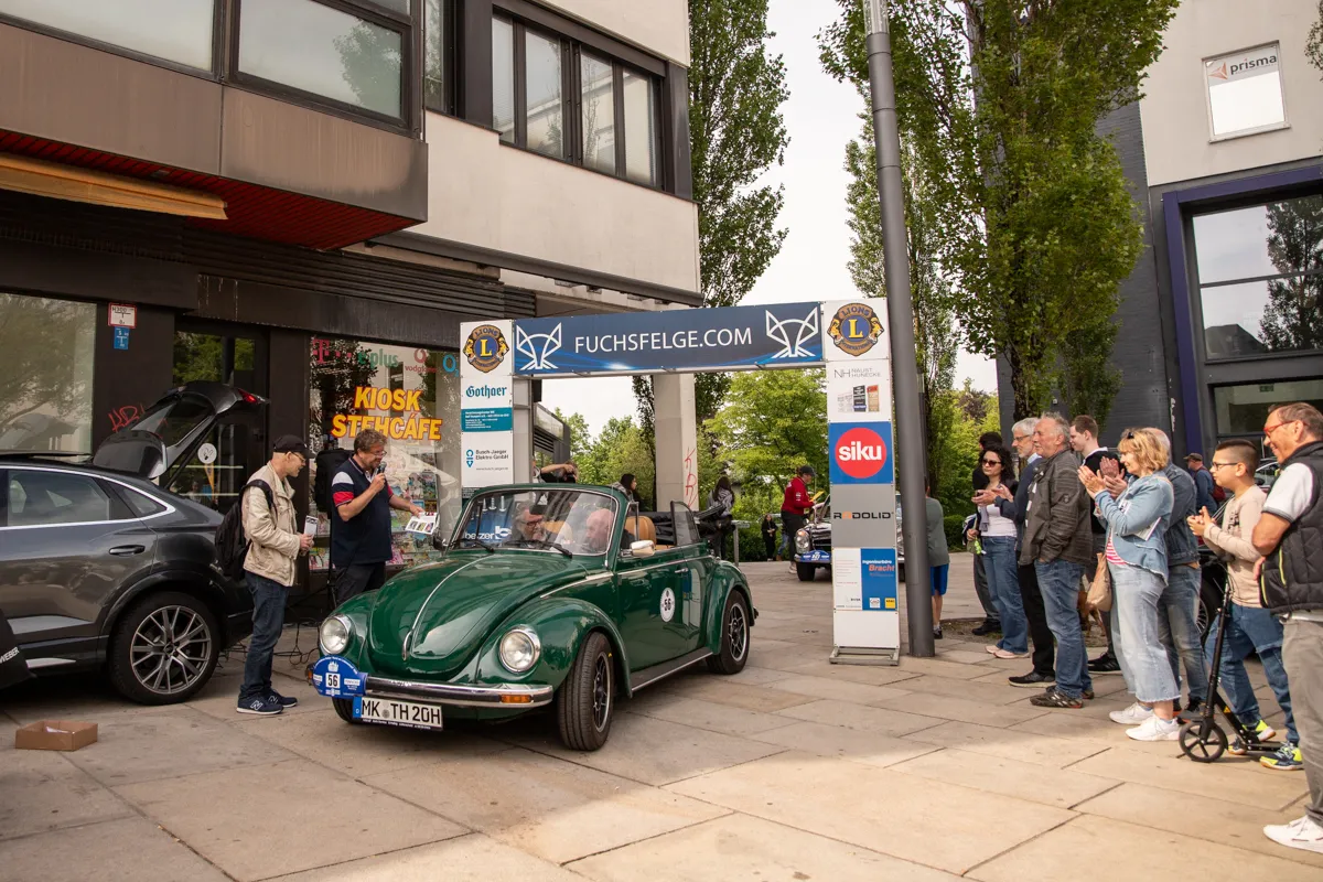 Ein dunkelgrüner VW Käfer steht im Mittelpunkt einer Menschenmenge vor Geschäften in einer Fußgängerzone. Im Hintergrund sind Werbebanner und ein grauer Tesla zu sehen.