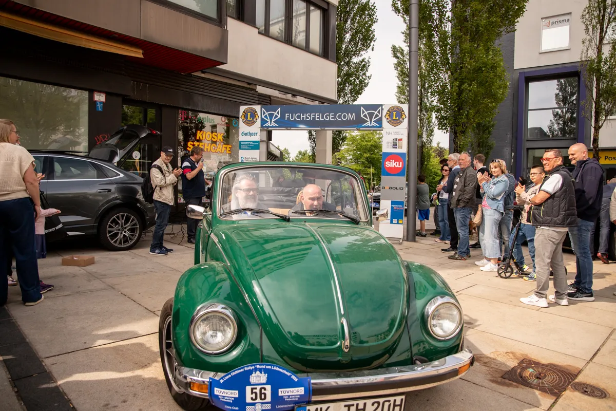 Grüner VW Käfer mit zwei Insassen fährt vor Zuschauern auf einer Straße durch die Lüdenscheider Innenstadt. Im Hintergrund Geschäfte und Passanten.