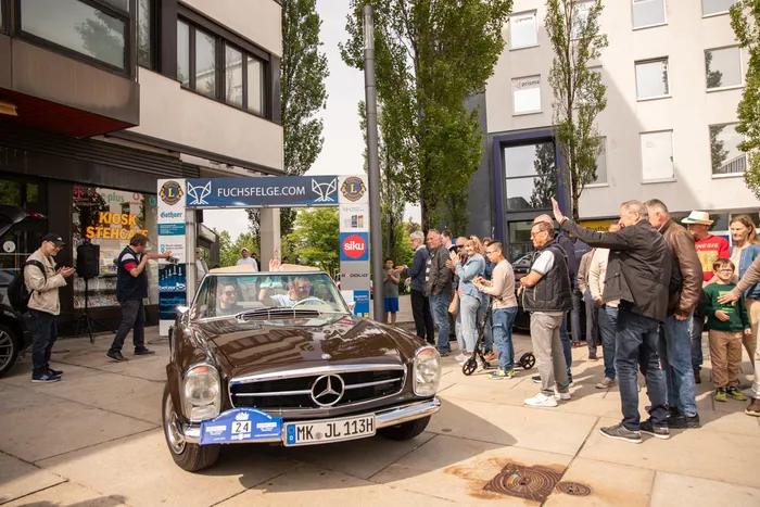 Ein brauner Oldtimer Mercedes steht auf einer Straße, umgeben von einer Menschenmenge, die zuschaut und fotografiert. Geschäfte säumen die Straße im Hintergrund.