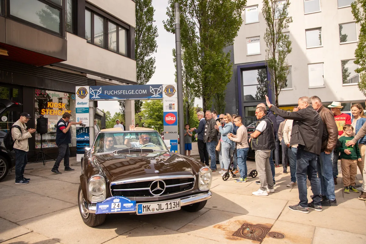 Ein brauner Oldtimer Mercedes steht auf einer Straße, umgeben von einer Menschenmenge, die zuschaut und fotografiert. Geschäfte säumen die Straße im Hintergrund.