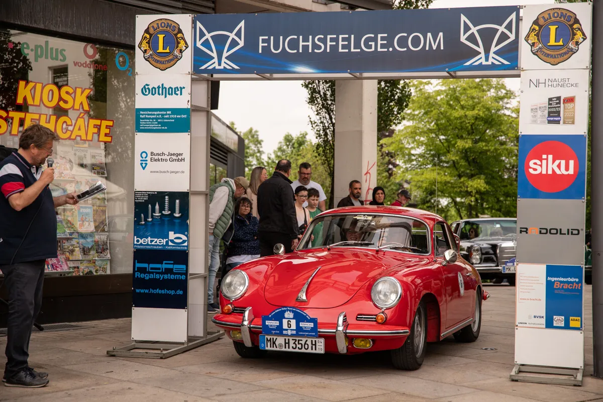 Roter Oldtimer steht vor einem Kiosk und Werbeaufstellern auf einer belebten Straße während eines Stadtfestes.