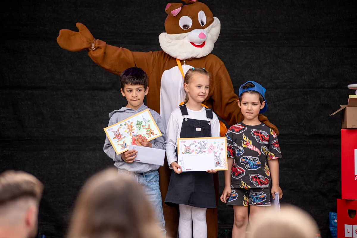 Drei Kinder stehen auf einer Bühne und halten jeweils ein Blatt Papier mit Zeichnungen. Im Hintergrund steht ein großes Eichhörnchen-Maskottchen.