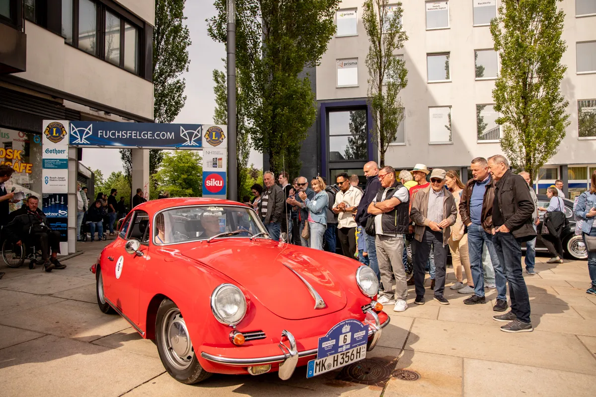 Roter Oldtimer-Sportwagen steht vor einer Gruppe von Zuschauern in einer Fußgängerzone. Im Hintergrund sind Geschäfte und Gebäude zu sehen.