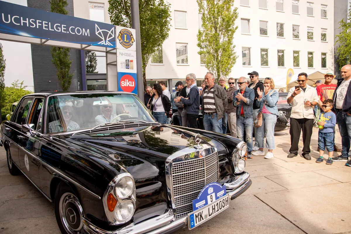 Schwarzer Oldtimer Mercedes steht vor Zuschauern und einem modernen Gebäude mit Werbeplakaten. Menschen beobachten die Veranstaltung „Frühling in der Lüdenscheider Innenstadt“.