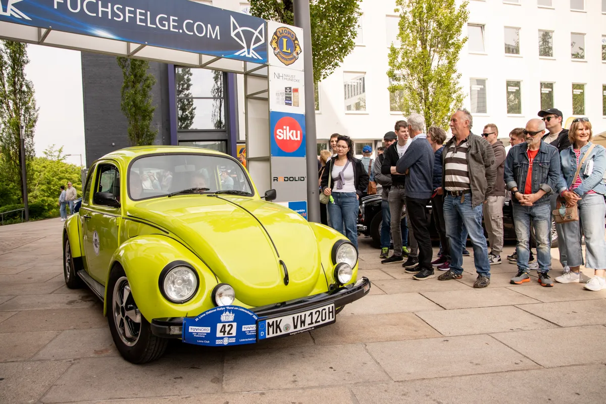 Gelber VW Käfer mit Startnummer 42 steht vor einer Gruppe von Zuschauern vor einem Gebäude mit Werbebanner.